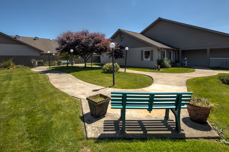Outdoor garden area at a senior living facility with a green bench on a concrete pad, two potted plants, a curved concrete walkway, several lamp posts, green grass, and a building with beige siding in the background under a clear blue sky.