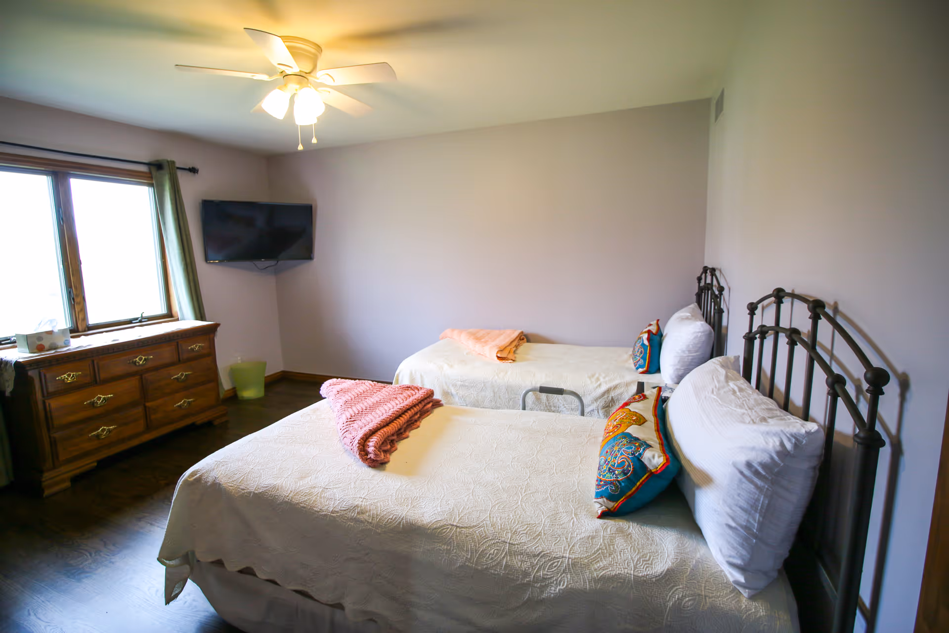 A bedroom with two single beds featuring metal headboards, white bedding, and colorful pillows. A wooden dresser is positioned under a window with green curtains. A ceiling fan with lights is mounted on the ceiling, and a flat-screen TV is mounted on the wall.
