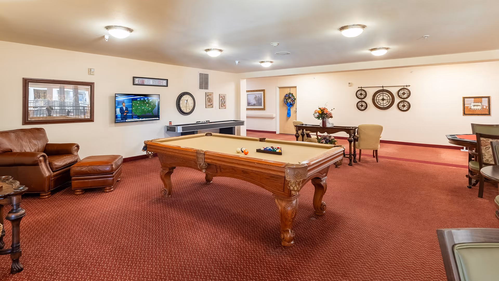A spacious recreational room with a pool table in the center, a leather armchair with an ottoman on the left, a wall-mounted TV showing a weather forecast, a shuffleboard table beneath the TV, and a table with chairs and floral decorations in the background. The room has red carpet flooring and cream-colored walls adorned with clocks, framed pictures, and a wreath on a door.