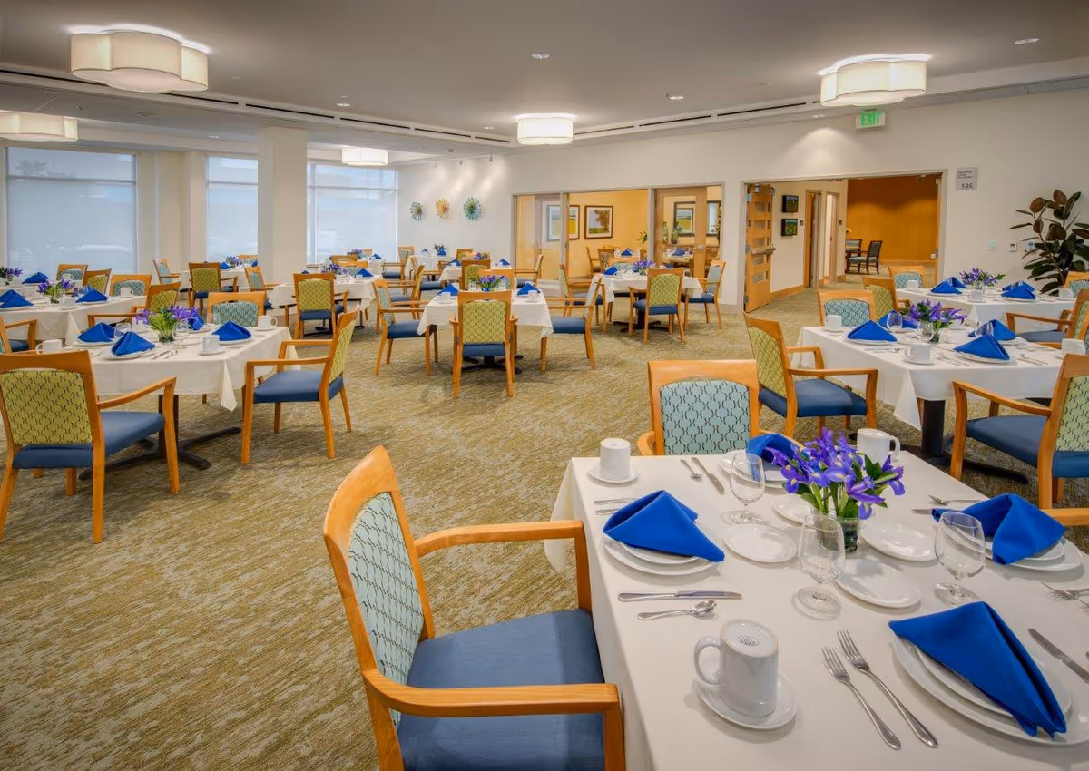 Well-lit dining room with multiple tables set with white linens, blue napkins, glassware and floral centerpieces.