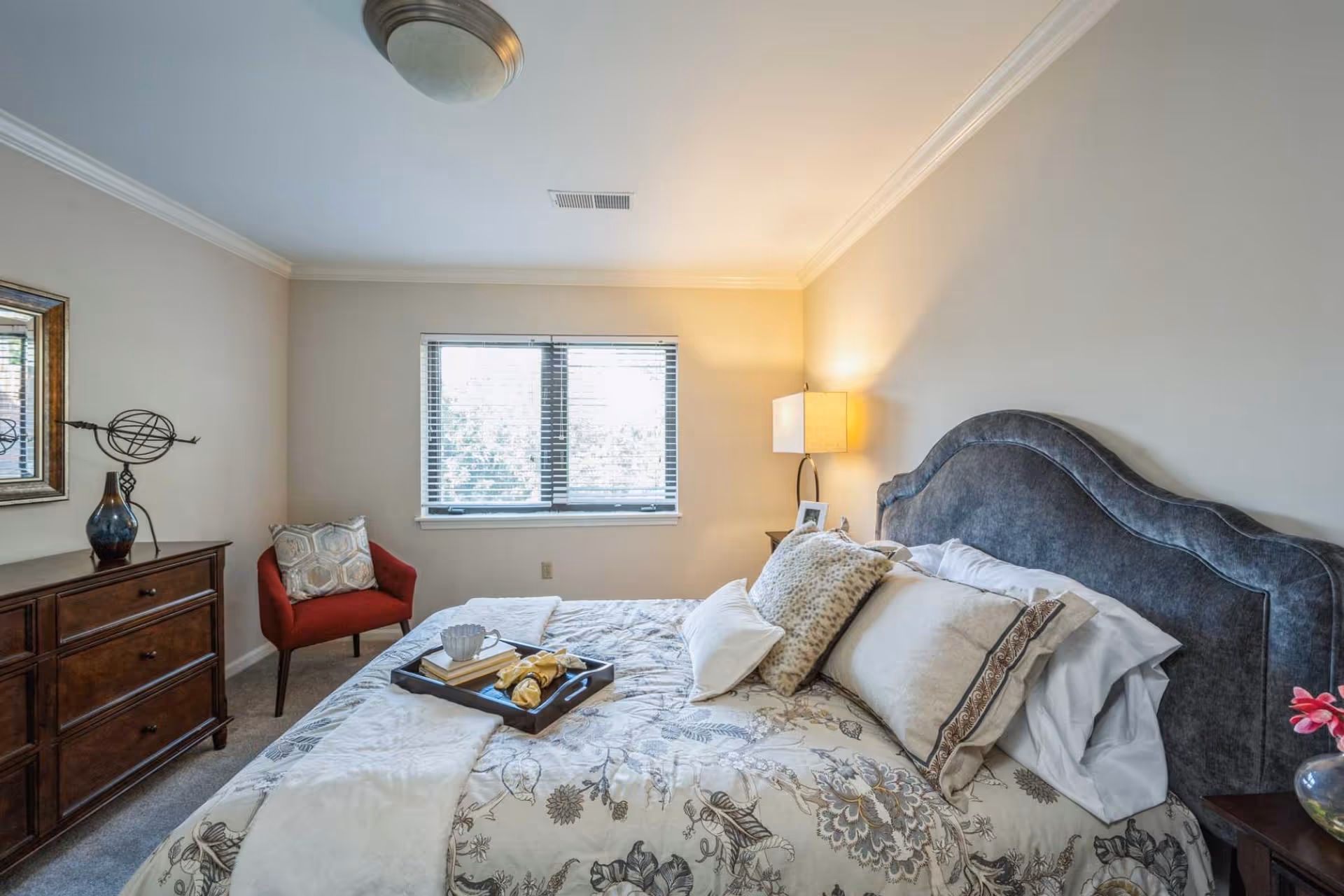 A cozy bedroom with a large bed featuring a floral patterned comforter and multiple pillows. There is a dark upholstered headboard, a wooden dresser with decorative items, a red armchair with a cushion, a window with blinds, and a bedside table with a lamp and a framed photo.