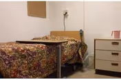A small bedroom with a single bed covered in a colorful patterned bedspread. Next to the bed is a small table on wheels and a three-drawer dresser with a pink pitcher on top. The walls are plain and light-colored, with an electrical outlet and a cord plugged in above the bed's headboard.