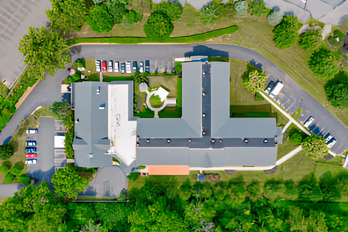 Aerial view of a senior living facility building with a T-shaped roof surrounded by green trees and parking lots with several cars parked.