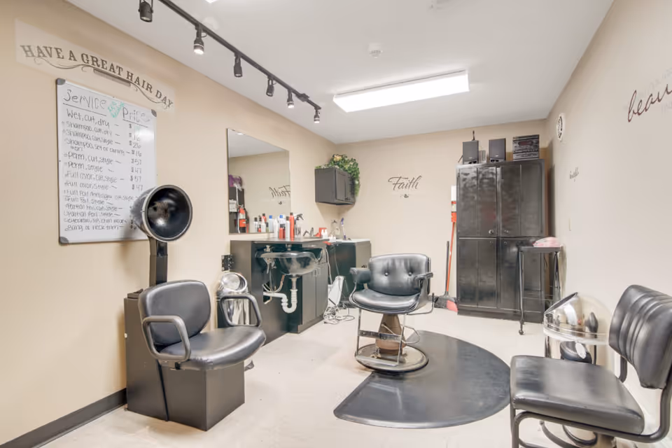 Interior of a hair salon room with black salon chairs, a hair dryer, a large mirror above a counter with hair products, and a black storage cabinet. The walls have decorative words like 'Faith' and 'Have a great hair day'. A whiteboard lists hair service prices.