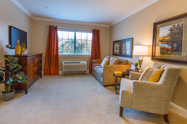 A cozy living room with beige walls and carpet, featuring a large window with orange curtains. There is a beige sofa with cushions, a beige armchair with a pillow, a wooden dresser with decorative items, a potted plant, a side table with a lamp, and framed artwork on the walls.