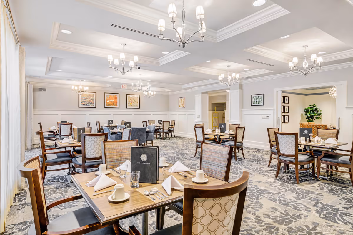 A bright and spacious dining room in an assisted living facility with multiple wooden tables and chairs arranged neatly. Each table is set with white napkins, cups, glasses, and silverware. The room features elegant chandeliers hanging from a coffered ceiling, patterned carpet flooring, and framed artwork on the walls. Large windows with curtains allow natural light to fill the space.