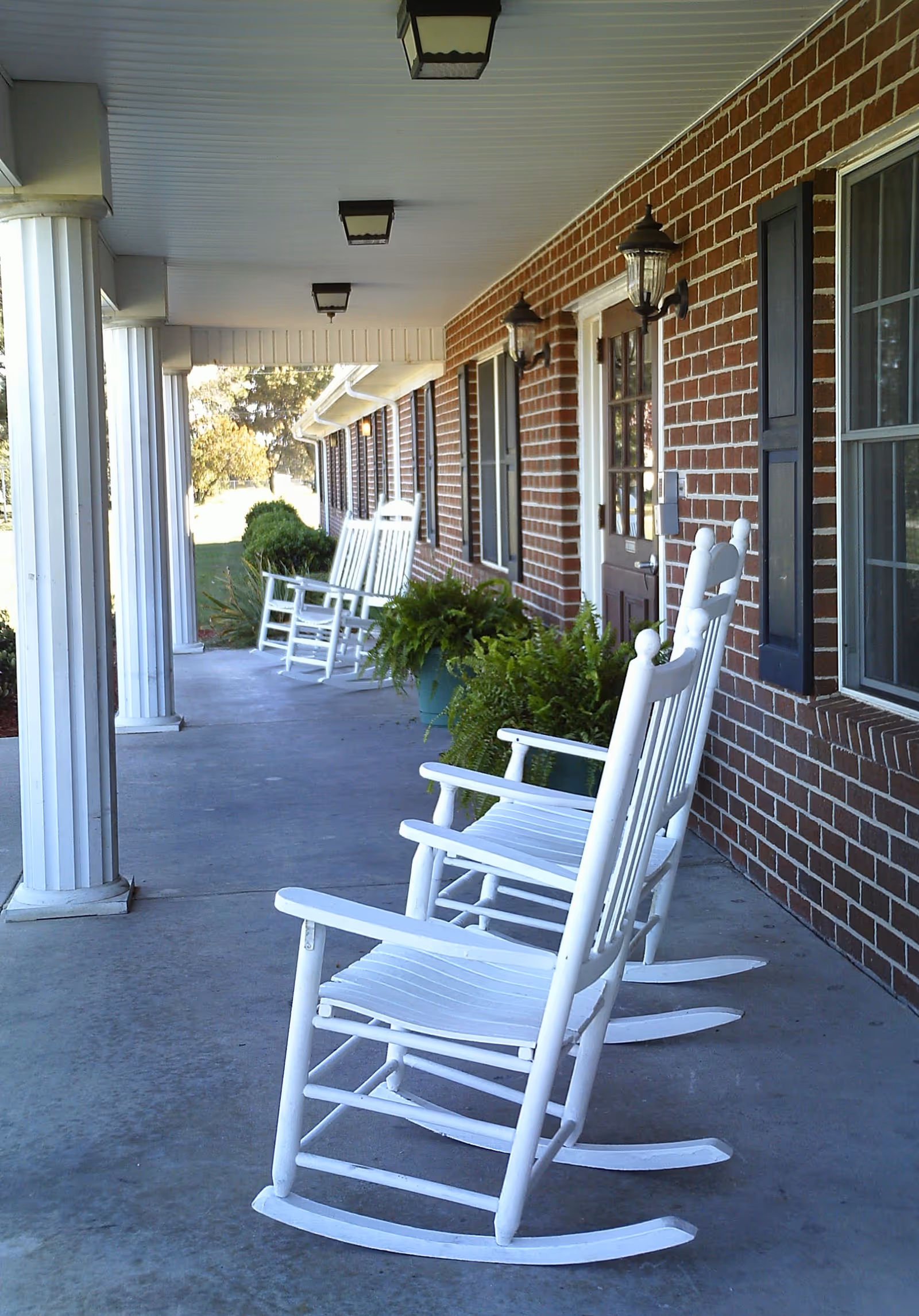 A covered brick front porch with white rocking chairs lined up and potted plants.