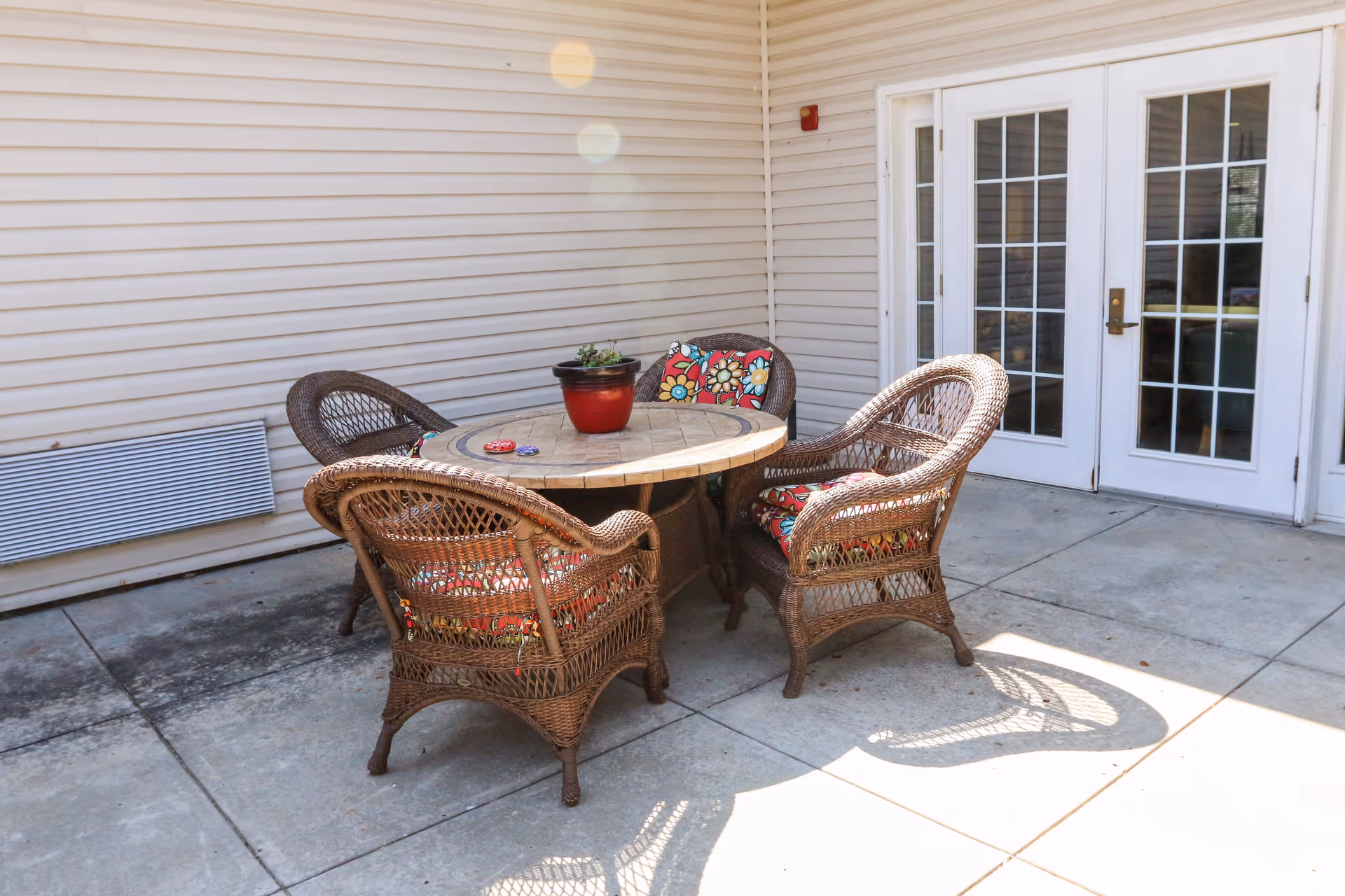 Outdoor patio area with a round table and four wicker chairs with colorful floral cushions. A small potted plant is on the table. The patio is adjacent to a building with beige siding and white French doors.
