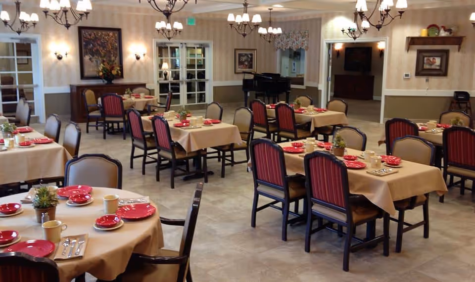 A dining room with multiple tables set for meals with red plates and beige tablecloths, chandeliers overhead and a piano in the background.