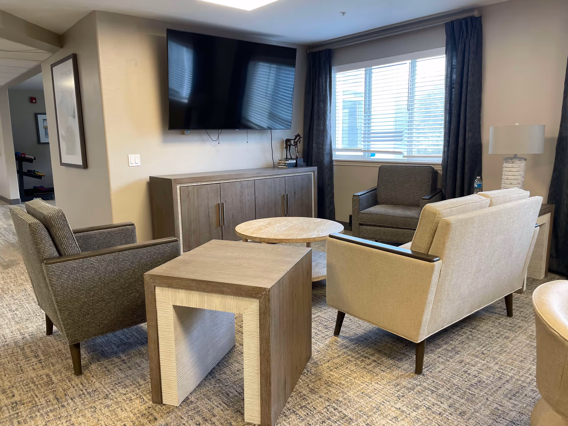A cozy living room area with a beige loveseat, two gray armchairs, a wooden side table, and a round coffee table. A large flat-screen TV is mounted on the wall above a wooden cabinet. A window with dark curtains allows natural light into the room, and a table lamp is placed on a side table next to the loveseat.