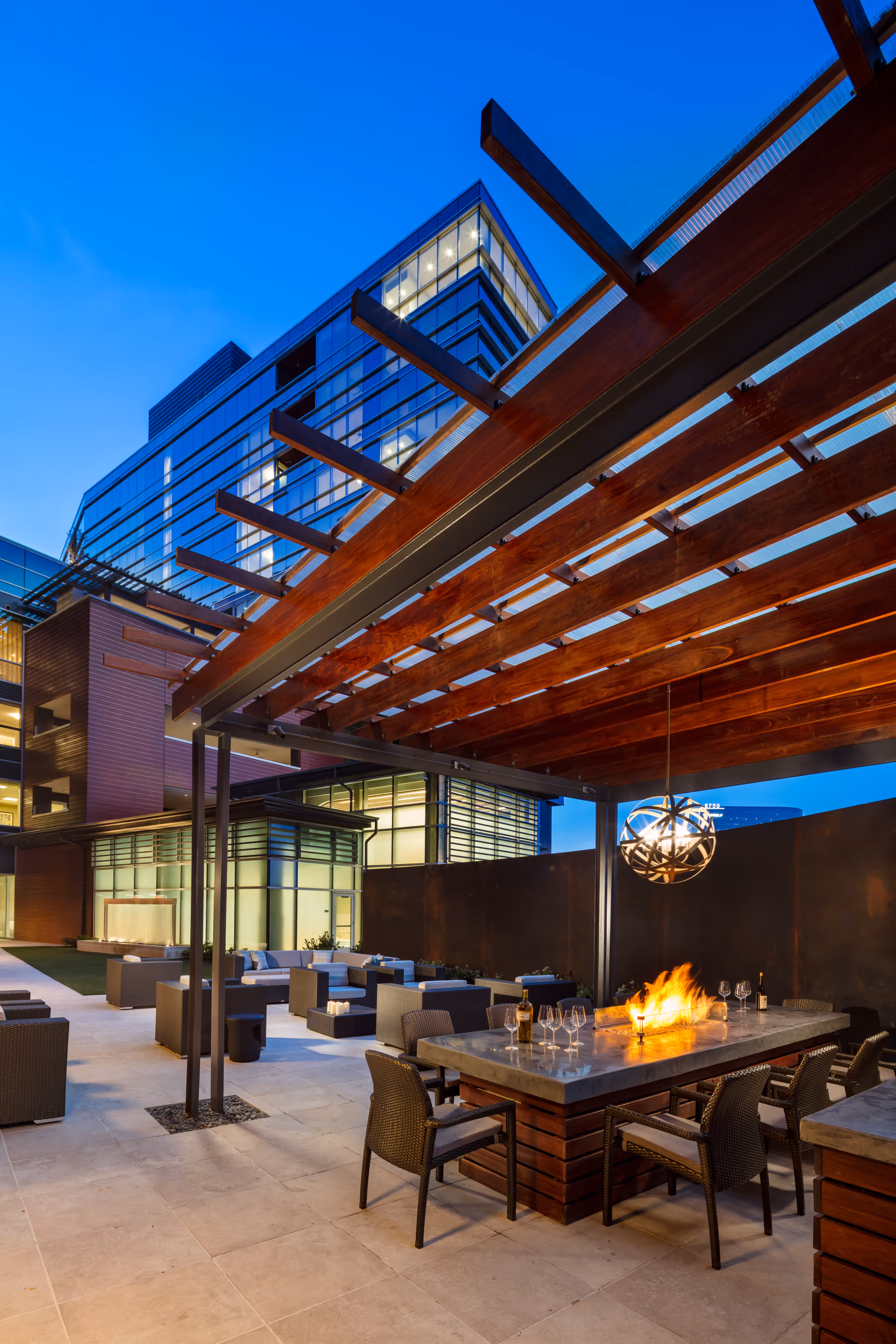 Outdoor patio area at dusk with modern seating arrangements, a fire pit table with chairs around it, and a wooden pergola overhead. A multi-story glass building is visible in the background with lights on inside.