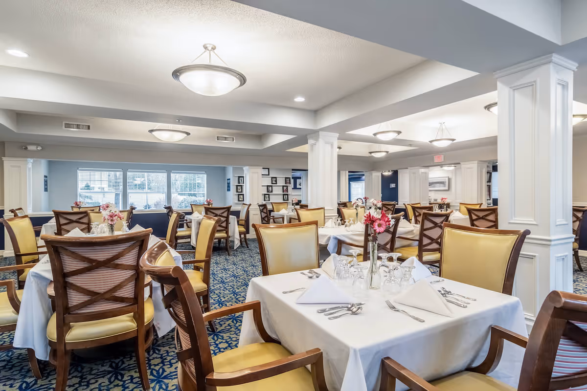 A bright and elegant dining room with multiple tables covered in white tablecloths, each set with folded napkins, glassware, and silverware. The chairs have wooden frames with yellow cushioned seats and backs. The room features white columns, blue patterned carpet, and large windows letting in natural light. Ceiling lights provide additional illumination.