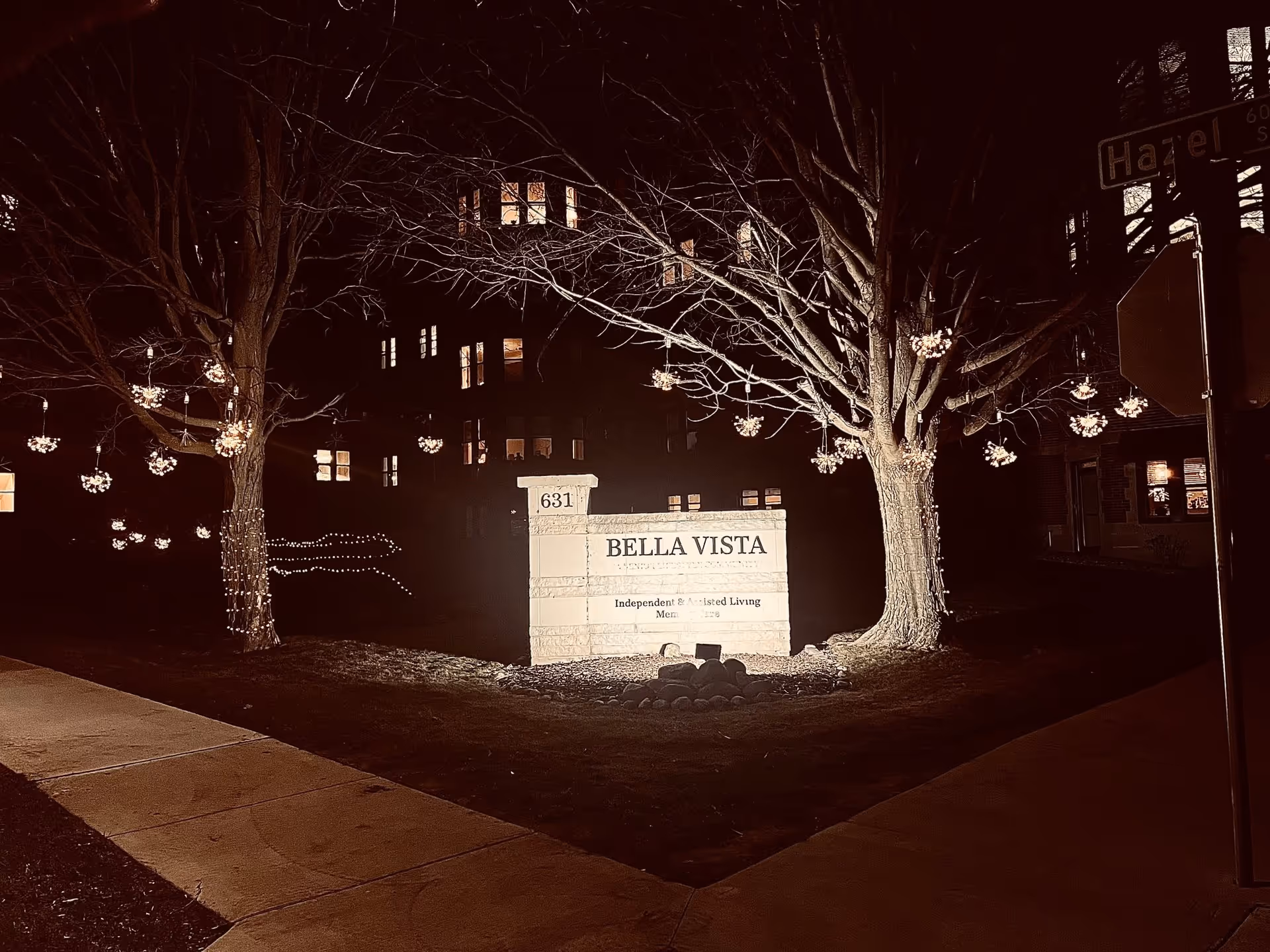 Nighttime view of the Bella Vista senior living facility entrance sign illuminated by lights. The sign is flanked by two trees decorated with hanging lights and string lights wrapped around the trunks. The building windows in the background are lit, and a street sign reading 'Hazel' is visible on the right side.