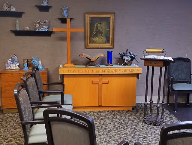Small chapel room with an altar bearing a wooden cross, a portrait of Jesus, a lectern, and rows of chairs.