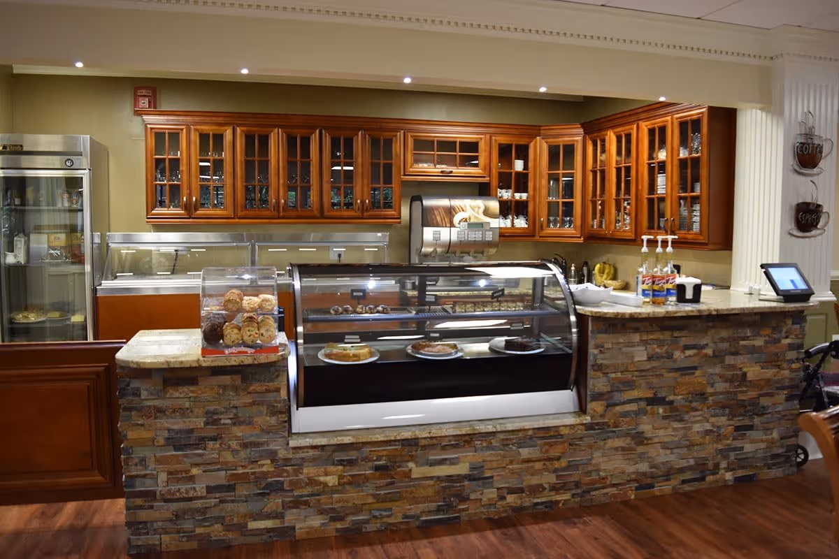 Interior view of a bakery or cafe counter area with a stone facade and glass display case containing cakes and pastries. Behind the counter are wooden cabinets with glass doors, a refrigerated display case, and a coffee machine mounted on the wall. The floor is wooden, and there are syrup bottles and a touchscreen register on the counter.