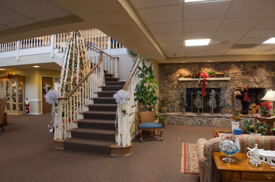 A cozy living room area in a senior living facility decorated for the holidays with a Christmas tree wrapped in lights and ribbons next to a carpeted staircase. The room features a stone fireplace adorned with festive garlands and a red bow, a small chair, a sofa with a patterned rug, and various holiday decorations including a small Santa figure and a bear statue wearing a scarf.