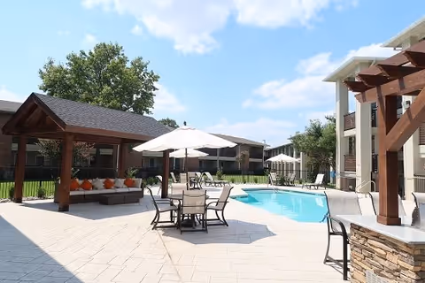 Outdoor pool area at MontClair Estates featuring a swimming pool, patio tables with umbrellas, lounge chairs, and a shaded seating area with cushions under a wooden pergola. The sky is clear with a few clouds and there are trees and buildings in the background.