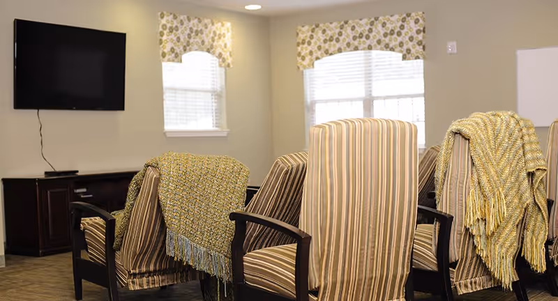 A cozy common area with several striped armchairs draped with knitted throws, two windows with patterned valances, a wall-mounted flat screen TV, and a dark wooden cabinet beneath the TV.