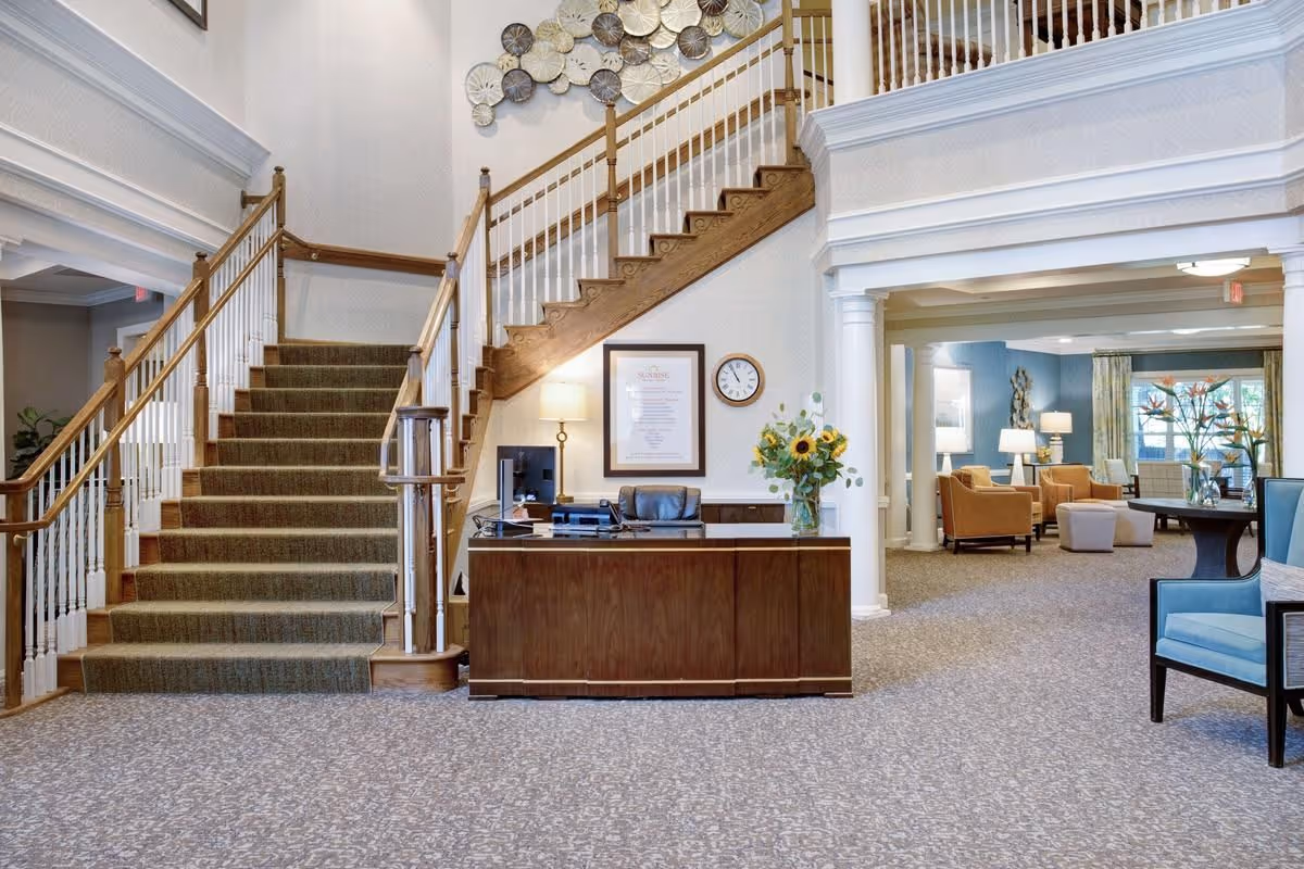 Reception desk in a bright senior living lobby with a central staircase, seating areas, and decorative wall art.