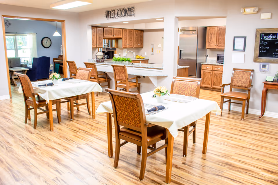 A bright dining area with wooden floors and tables covered with white tablecloths, each set with napkins and small flower arrangements. In the background, there is a kitchen with wooden cabinets, a stainless steel refrigerator, and a countertop with bar stools. A 'WELCOME' sign is displayed above the kitchen pass-through window. To the left, a living room area with chairs and a clock on the wall is visible.