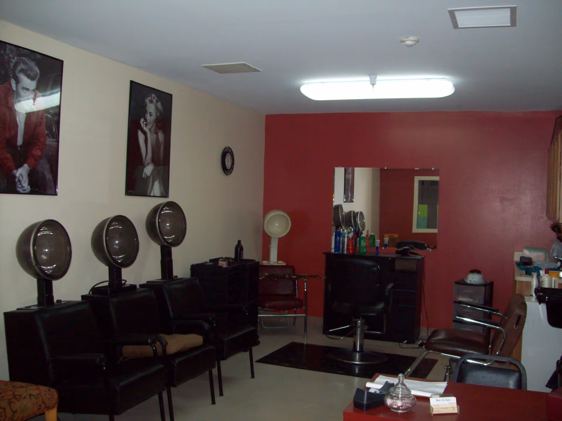 Interior of a hair salon room with three black hair drying chairs on the left, a styling chair in front of a mirror on a red accent wall, various hair care products on a counter, and two black and white celebrity posters on the wall.