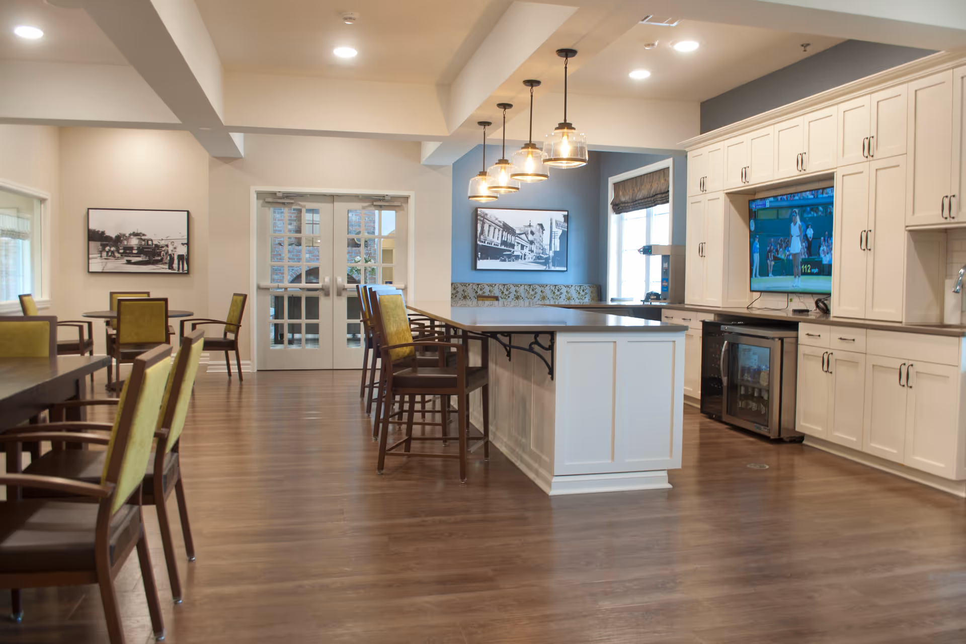A spacious and well-lit common area with a kitchen and dining space. The kitchen features white cabinetry, a large island with bar stools, pendant lights hanging above, and a mounted television. The dining area has several tables and chairs with green upholstery. There are large windows and double glass doors letting in natural light, and framed black and white photographs on the walls.