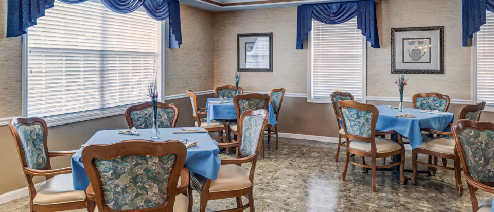 Bright dining room with several small round tables covered in blue tablecloths and wooden chairs with floral upholstery.