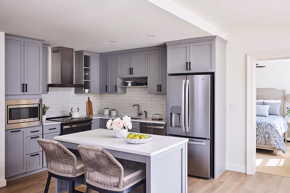 Bright modern kitchen with gray cabinets, a white island with two woven stools and a bowl of apples, stainless steel appliances, and a view into a bedroom.
