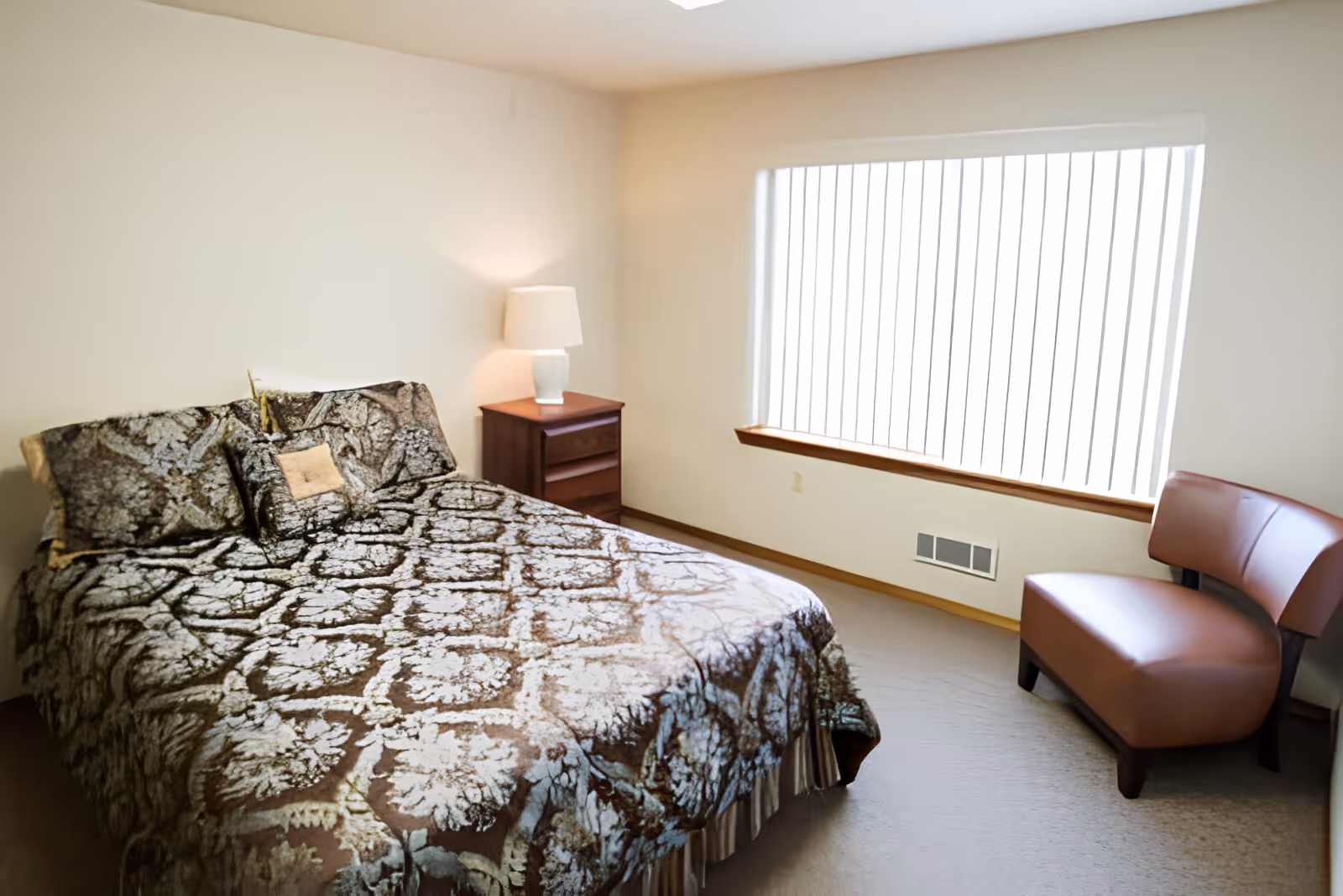 Furnished bedroom with a patterned bed, nightstand and lamp, large window with vertical blinds, and a chair.