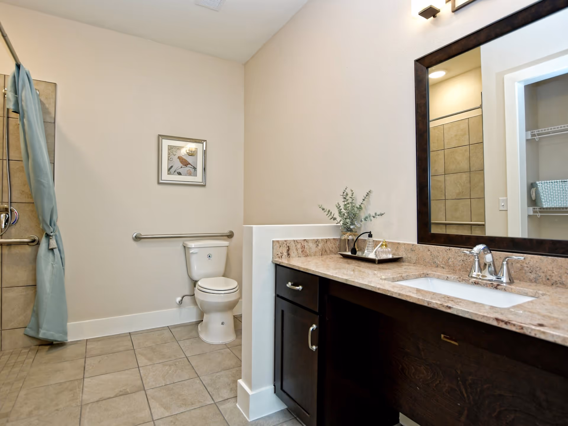 Bright accessible bathroom featuring a toilet with grab bars, a marble-top vanity and sink with large mirror, and a tiled walk-in shower with curtain.
