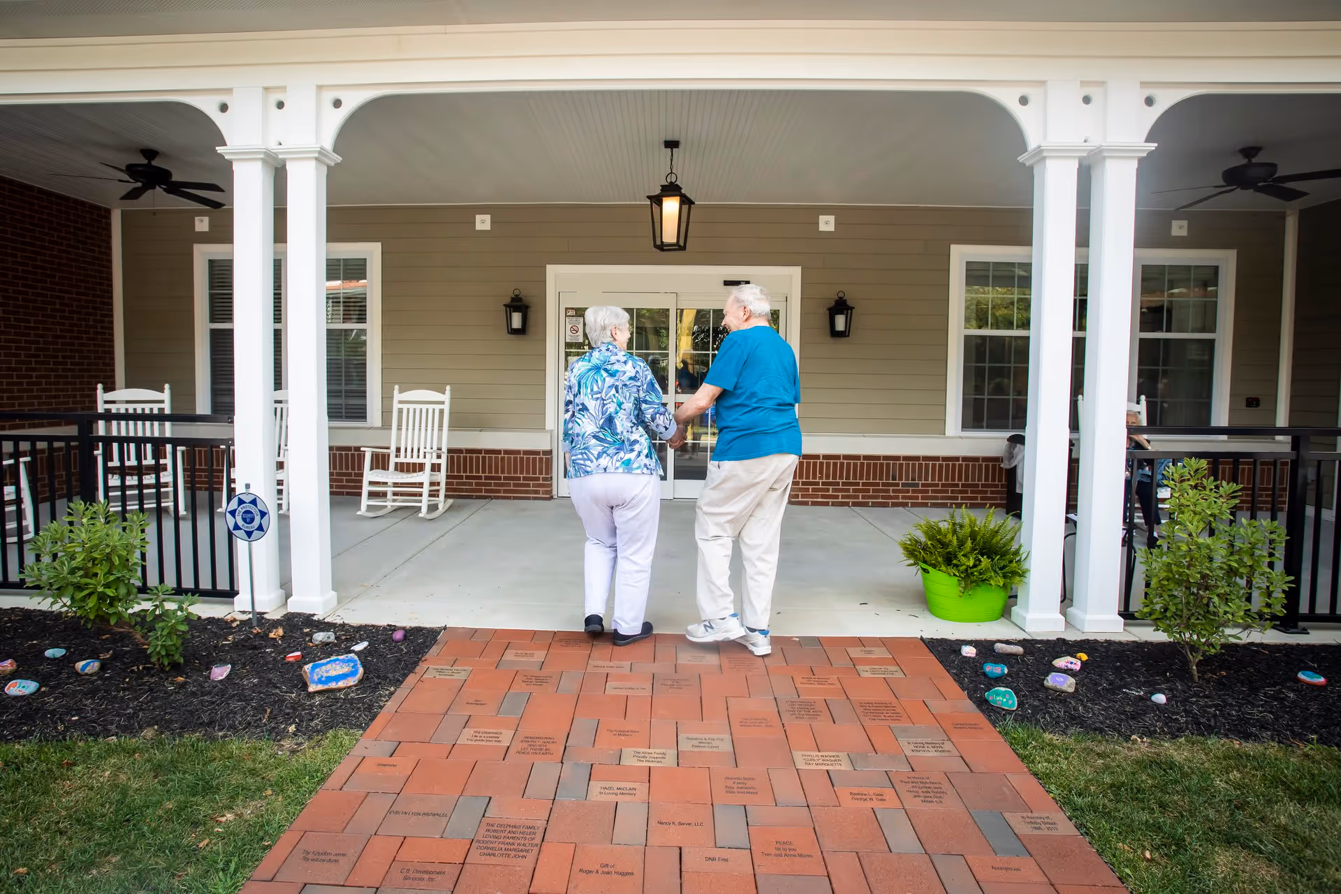 An elderly couple holding hands and walking towards the entrance of a building with a covered porch. The porch has white columns, rocking chairs, and ceiling fans. The walkway is made of engraved bricks, and there are small plants and painted rocks on either side of the path.