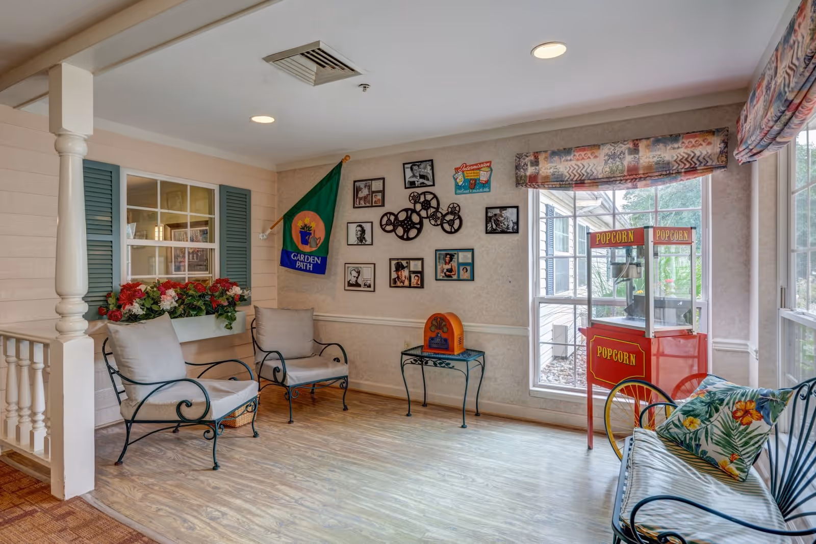 A cozy sitting area with two cushioned chairs and a cushioned bench with floral pillows. There is a red popcorn machine near a large window with patterned valances. The walls are decorated with framed pictures, a green flag with the text 'Garden Path,' and a decorative film reel wall art. A small table holds a vintage-style radio. The floor is wood, and the space is well-lit with ceiling lights.