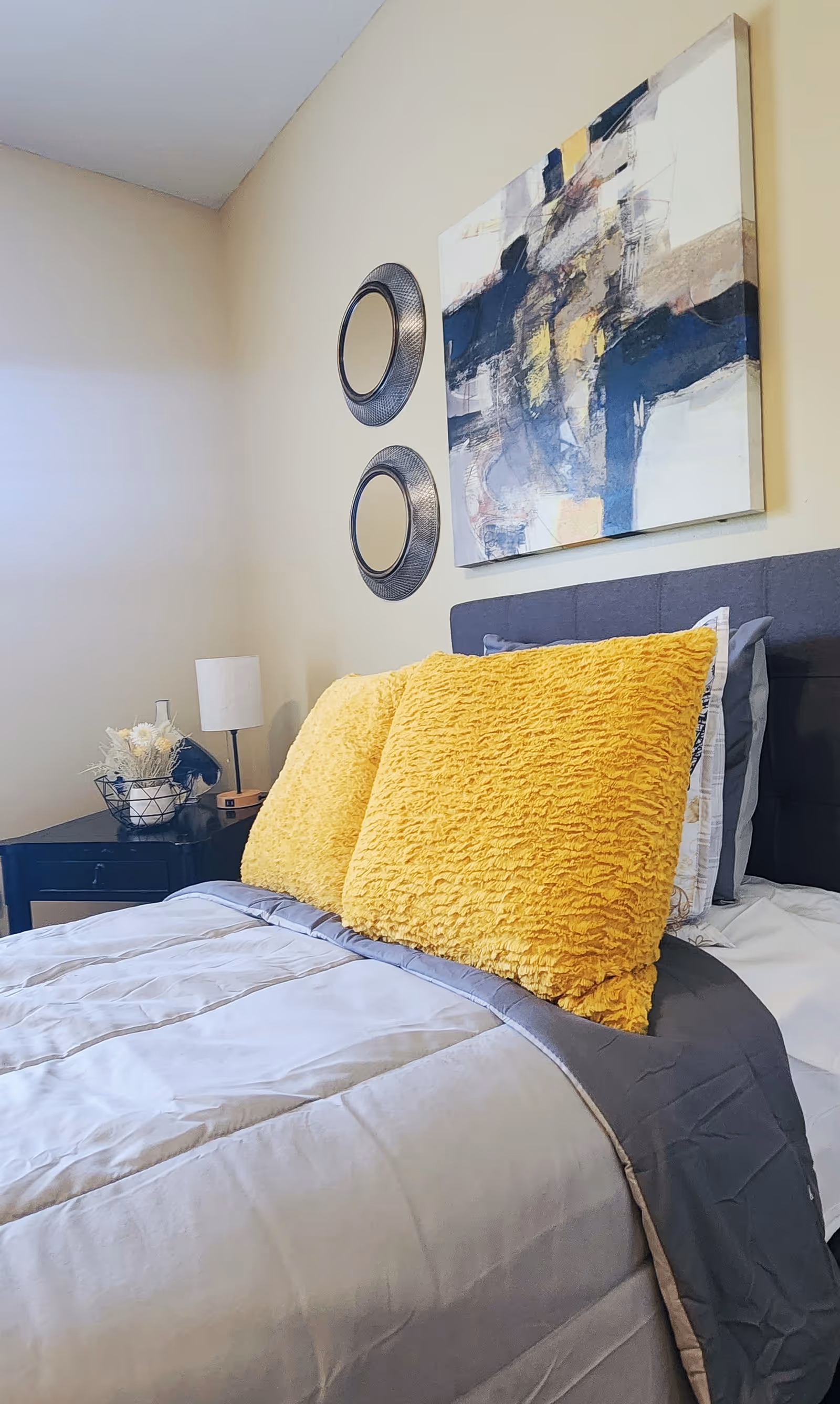 A neatly made bed with two large yellow textured pillows and gray bedding in a bedroom. Above the bed, there is an abstract painting and two round decorative mirrors on a beige wall. A black nightstand with a lamp and a decorative basket with flowers is next to the bed.