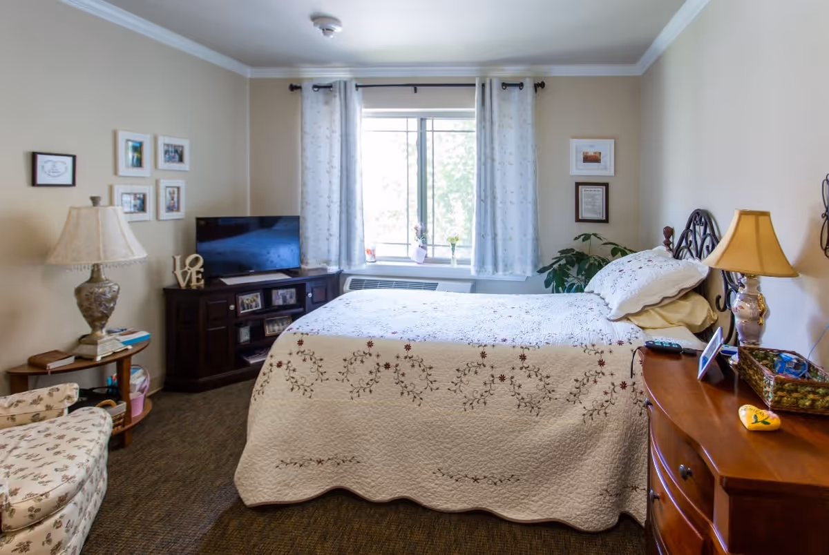 A cozy bedroom in an assisted living facility featuring a bed with a floral quilt, a wooden nightstand with a lamp and decorative items, a TV on a dark wooden cabinet, a cushioned armchair with floral upholstery, framed pictures on the walls, and a window with white curtains letting in natural light.