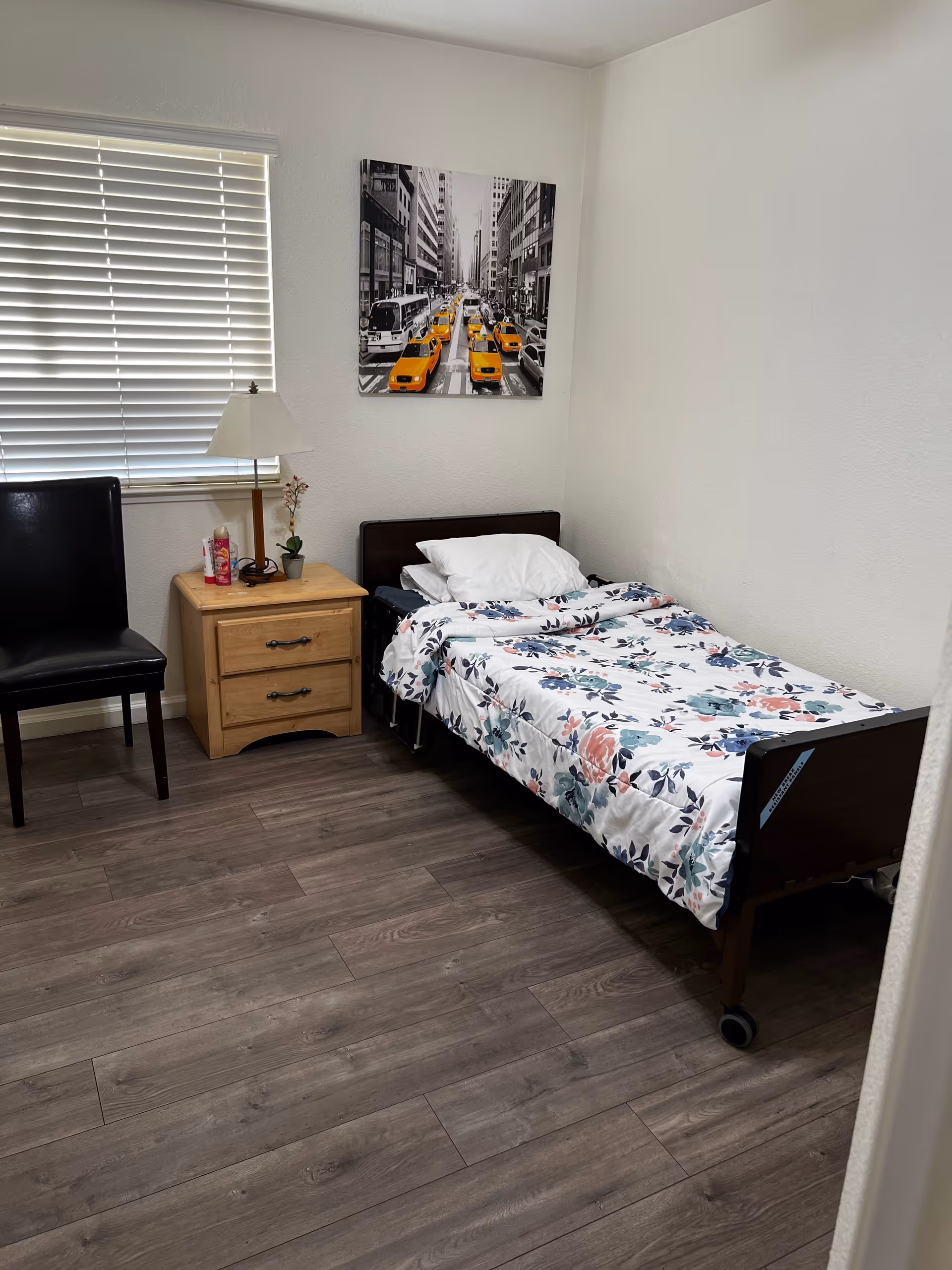 A small bedroom with a single bed covered in a floral patterned blanket. Next to the bed is a wooden nightstand with two drawers, a lamp, a small plant, and a bottle of lotion. A black chair is positioned near a window with closed blinds. A black and white photo with yellow taxis is hung on the wall above the bed.