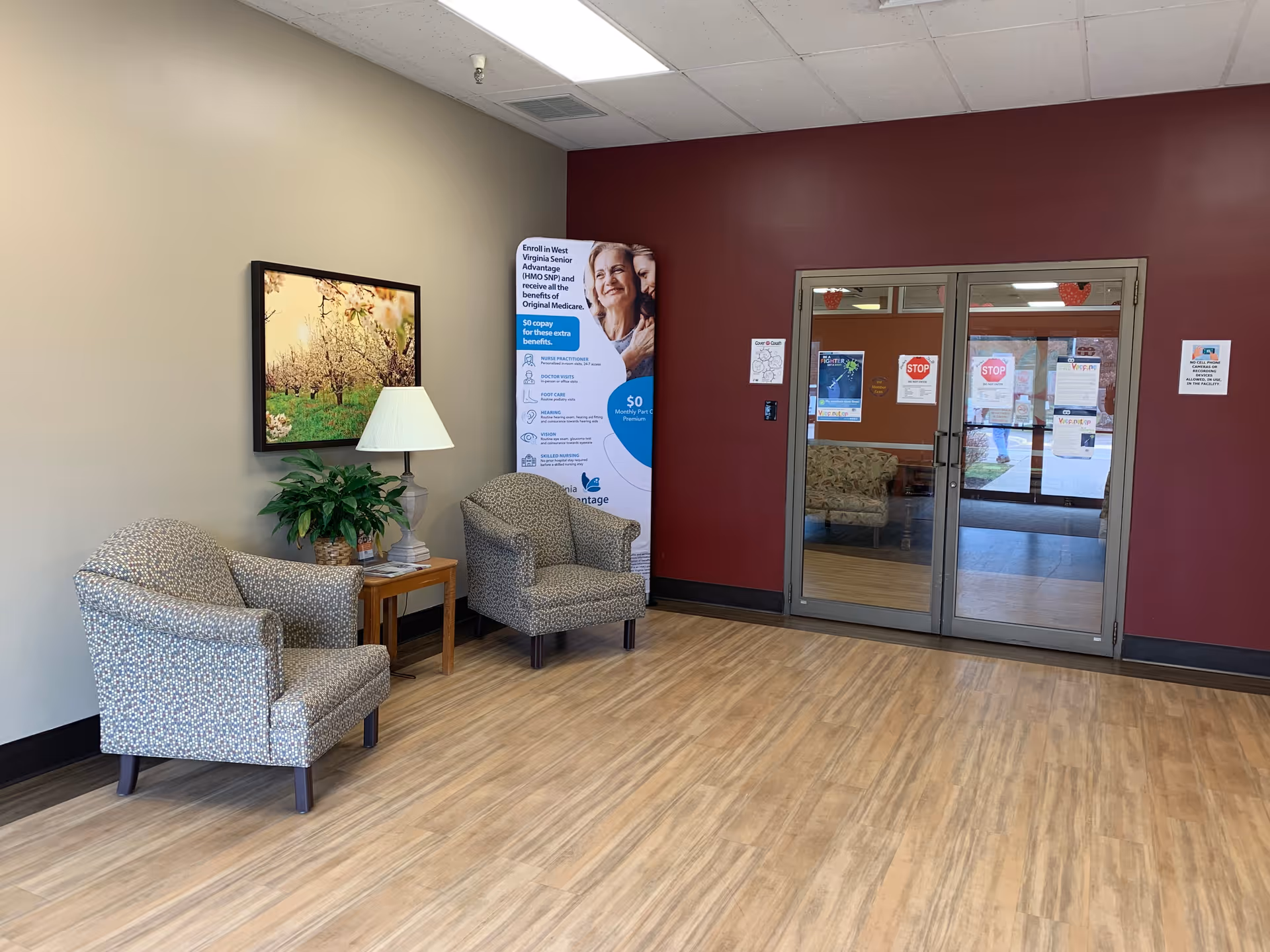 A waiting area with two patterned armchairs and a wooden side table with a lamp and a potted plant. A framed picture of a blossoming tree hangs on the beige wall. There is a maroon accent wall with double glass doors leading to another room. A standing informational banner about West Virginia Senior Advantage is placed near the corner.