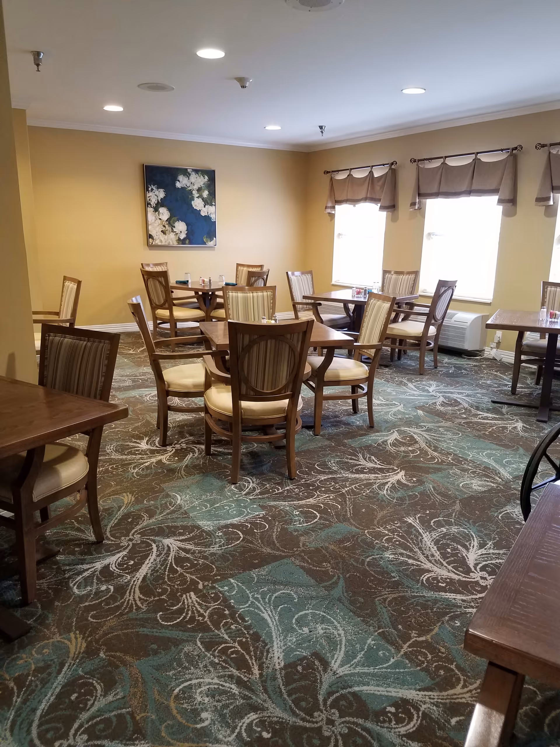 A bright dining room with multiple wooden tables and upholstered chairs on a patterned carpet beneath windows and wall art.