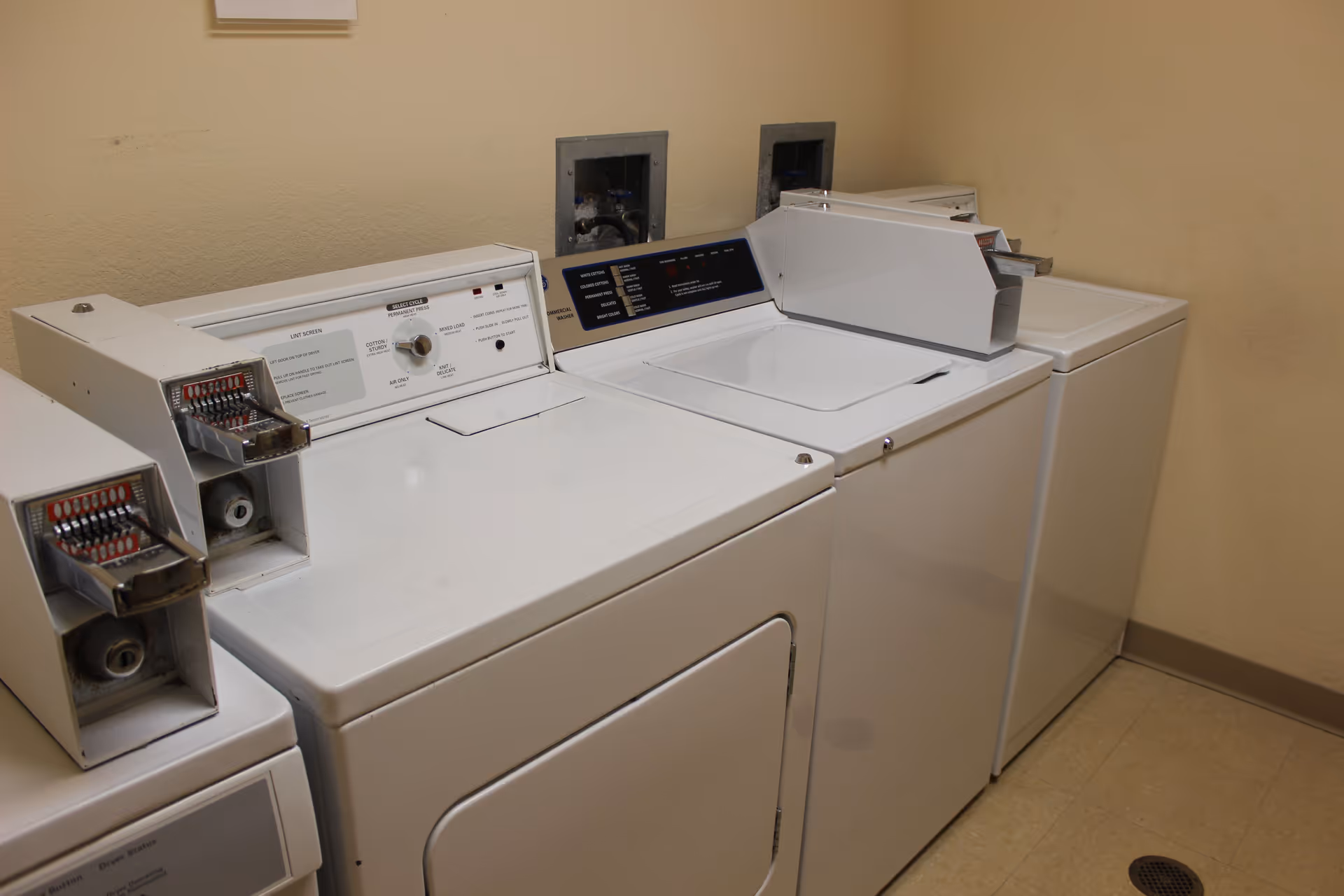 A laundry room with three white coin-operated washing machines and dryers lined up against a beige wall with a tiled floor.