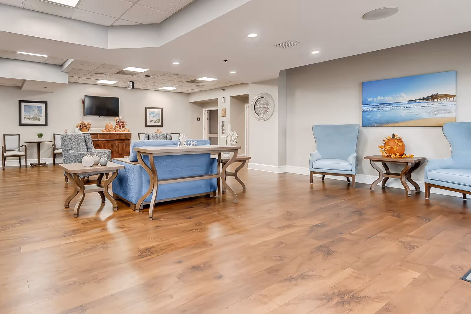 A spacious senior living facility common area with wooden flooring, light blue armchairs, a blue sofa, wooden tables, and wall decorations including a large beach scene painting and framed pictures. The room is well-lit with ceiling lights and has a welcoming, cozy atmosphere.