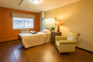A cozy nursing facility bedroom with a single bed covered in a beige quilt, a wooden nightstand with a lamp and a plant, a comfortable beige armchair, a window with blinds and a valance, and wooden flooring.