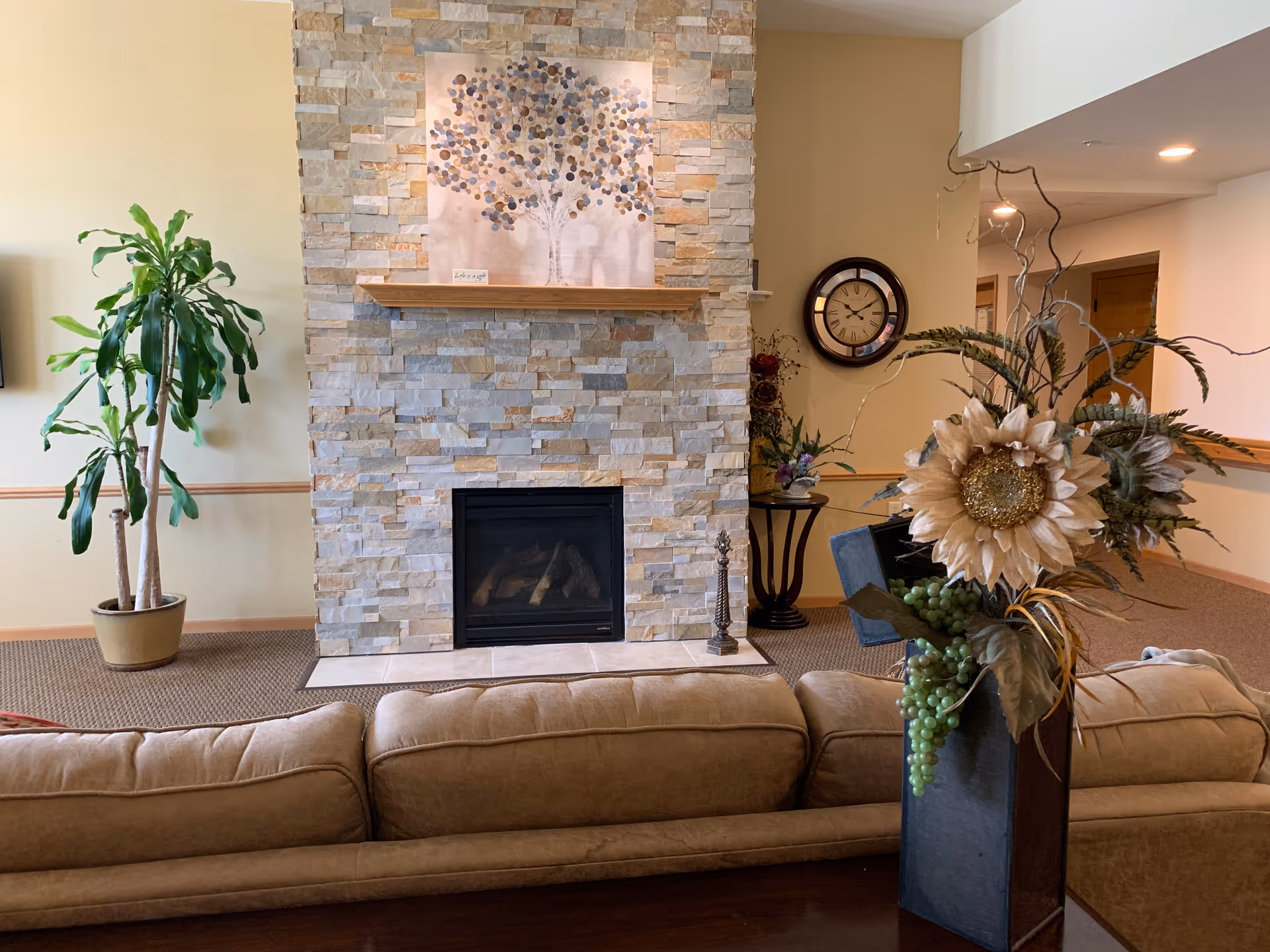 A cozy living room area with a stone fireplace in the center. Above the fireplace is a decorative painting of a tree with circular leaves. To the left of the fireplace is a tall potted plant. In front of the fireplace is a beige sofa, and to the right is a tall vase with artificial flowers and greenery. A round wall clock is visible on the wall in the background.