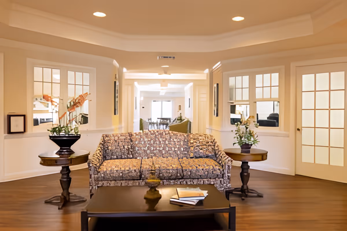 A cozy living room area with a patterned sofa centered between two round wooden side tables, each holding a potted plant. In front of the sofa is a dark wooden coffee table with some books on it. The room has warm lighting, wooden flooring, and white walls with windows and a door featuring multiple glass panes.