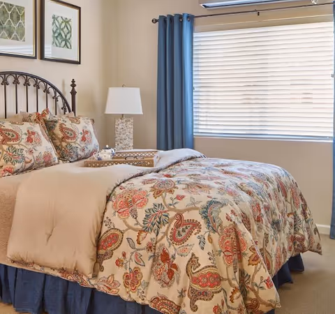 A decorated bedroom with a patterned bedspread, matching pillows, a nightstand lamp, and a window with blinds and blue curtains.
