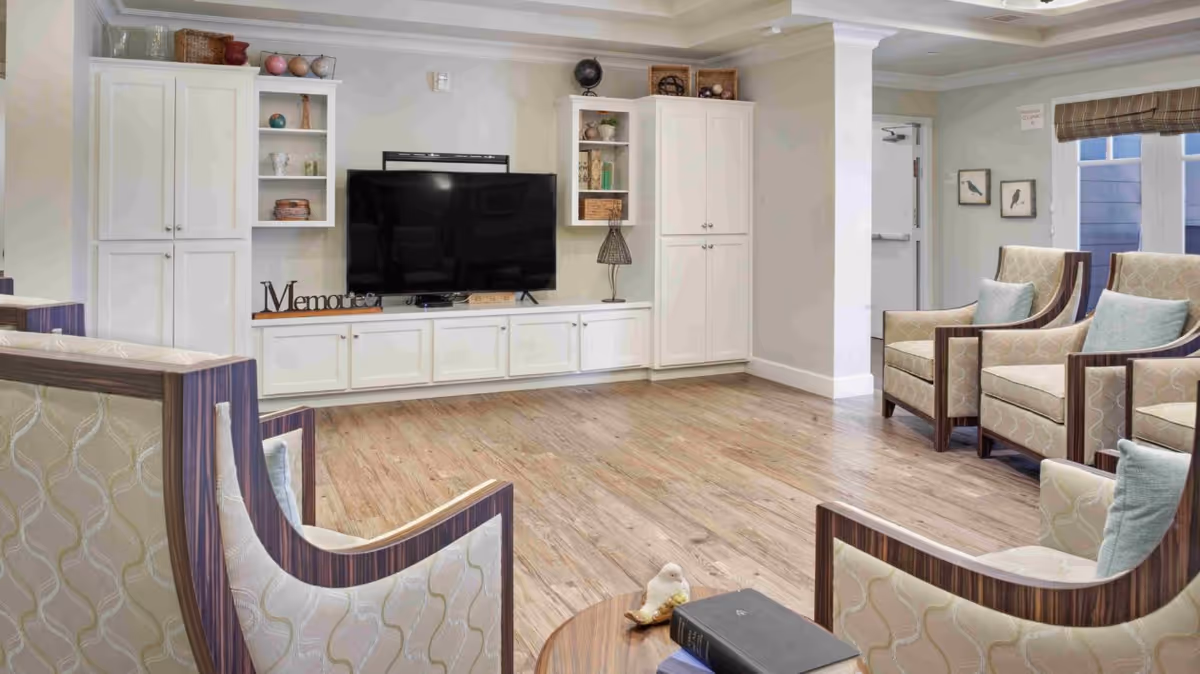 A cozy living room area with wooden flooring, several upholstered armchairs with wooden frames and light-colored patterned fabric, a large flat-screen TV mounted on a white built-in cabinet with shelves displaying decorative items, and a small round table with books and a bird figurine in the foreground.