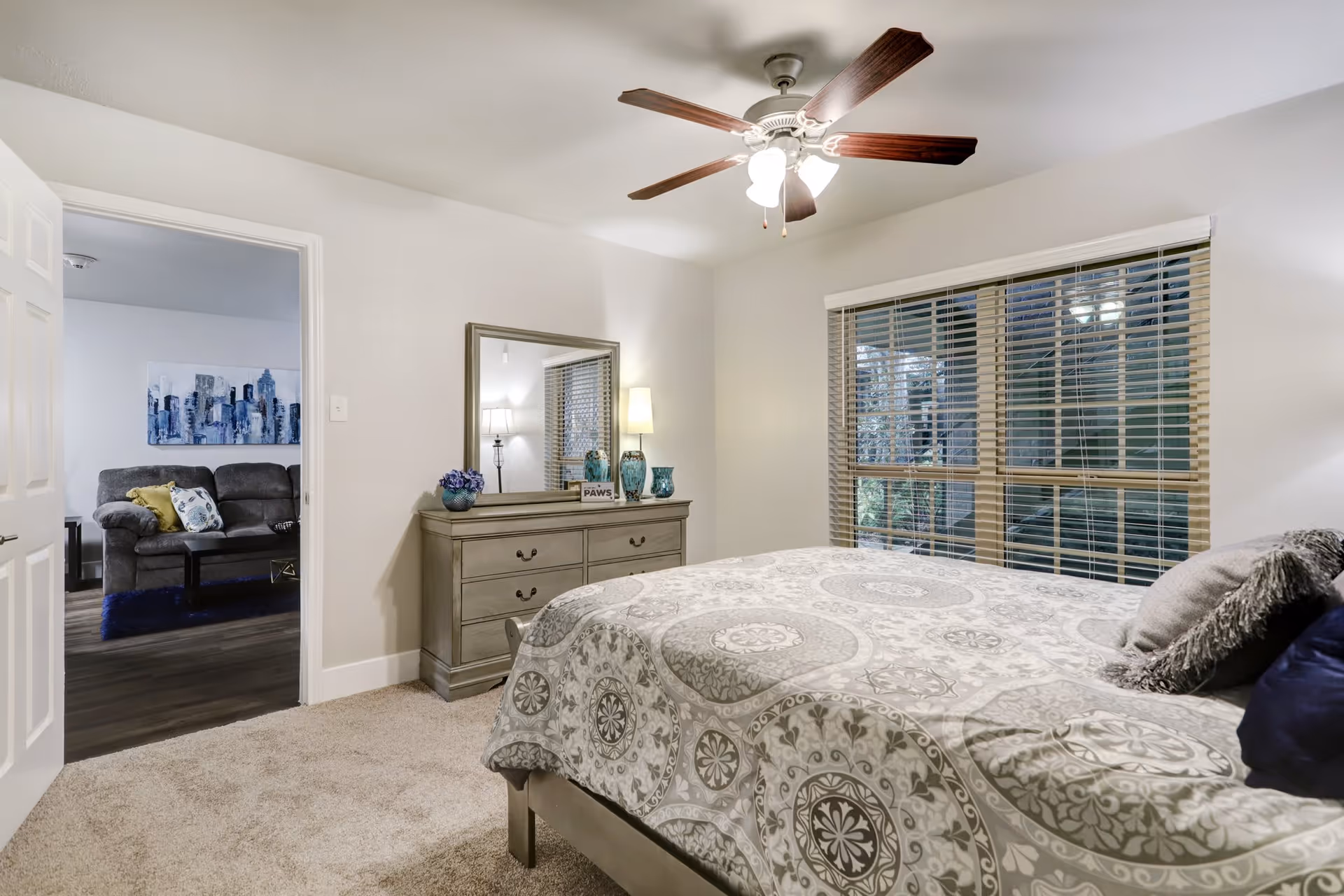 A cozy bedroom with a patterned bedspread on a bed, a dresser with a large mirror, decorative vases, and a lamp. A ceiling fan with lights is mounted on the ceiling. The room has a window with blinds showing an outdoor view. An open door leads to a living room area with a gray couch and a cityscape painting on the wall.