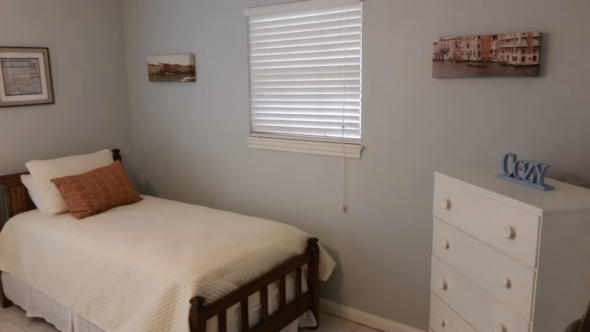 A simple bedroom with a single wooden bed covered with a white quilt and two pillows, one white and one brown with a pattern. There is a white chest of drawers with a decorative item spelling 'Cozy' on top. The walls are light gray with three framed pictures hanging. A window with white blinds is centered on the wall above the bed.