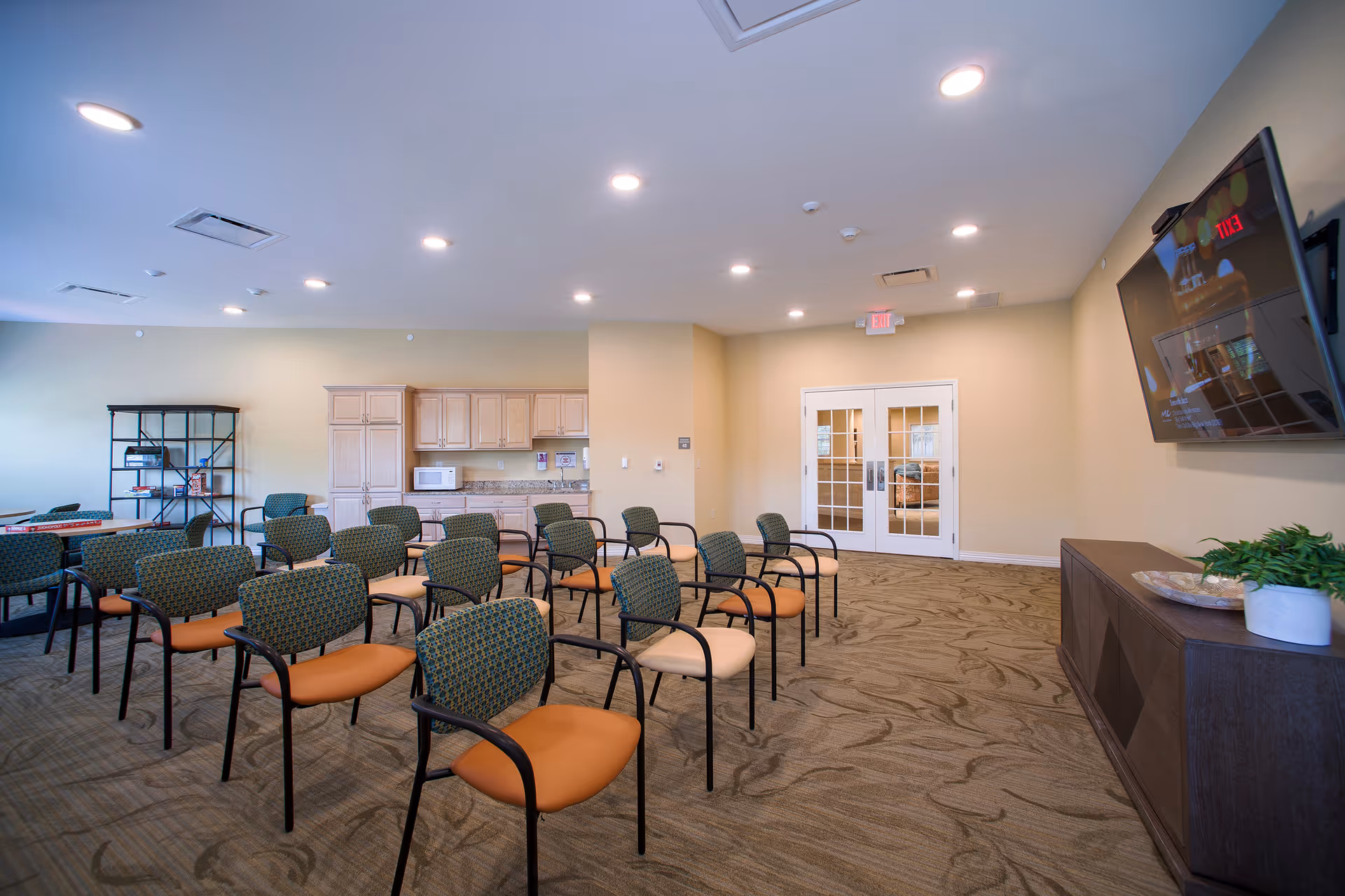 A spacious room with multiple rows of chairs arranged facing a wall-mounted television. The room has beige walls, a patterned carpet, and recessed ceiling lights. On one side, there is a kitchenette area with cabinets, a microwave, and a countertop. Double glass doors are visible at the far end of the room.