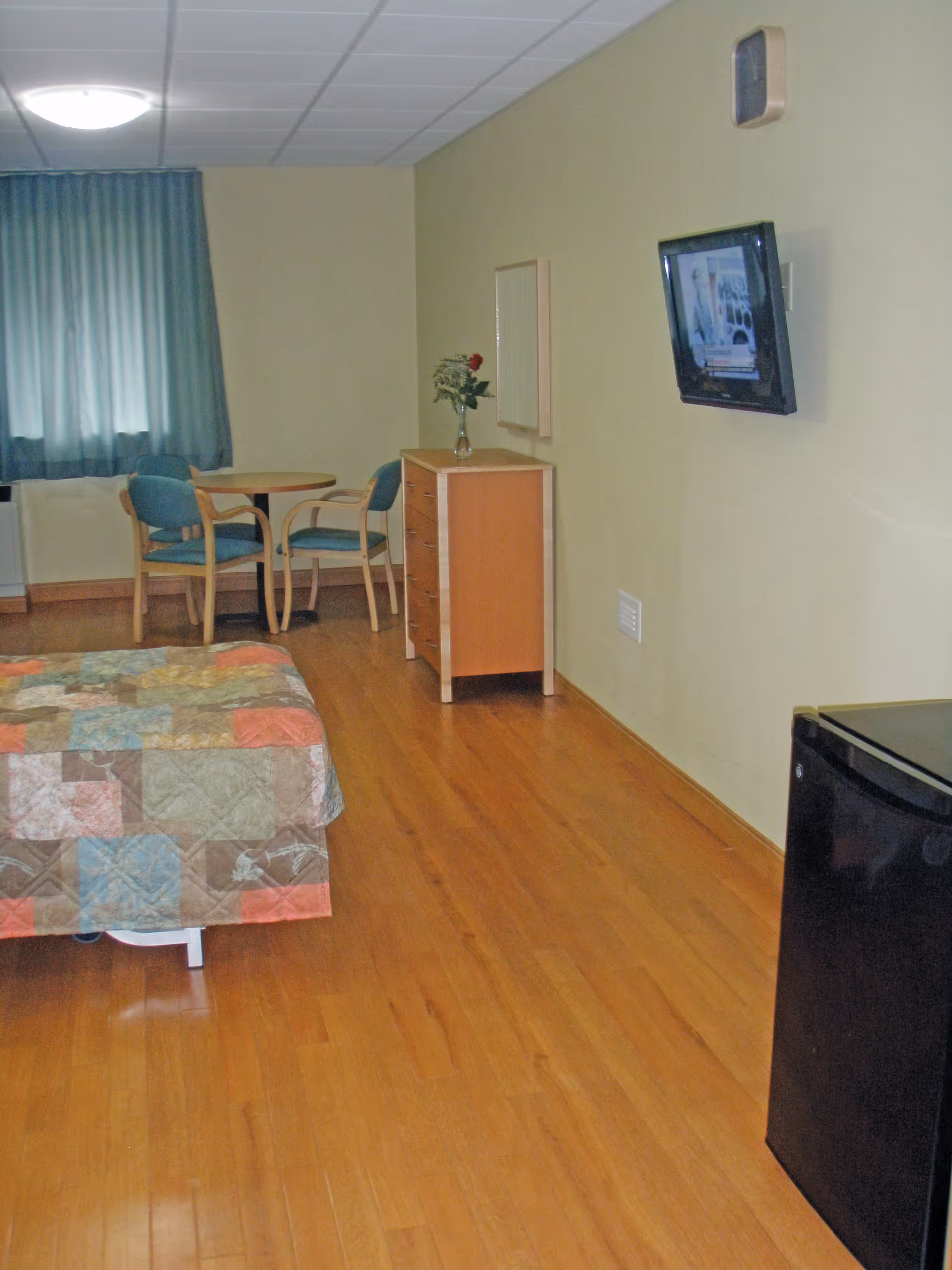 A senior living facility room with wooden flooring, a bed covered with a patchwork quilt, a small round table with two chairs near a window with green curtains, a wooden dresser with a vase of flowers on top, a wall-mounted TV, and a small black refrigerator.
