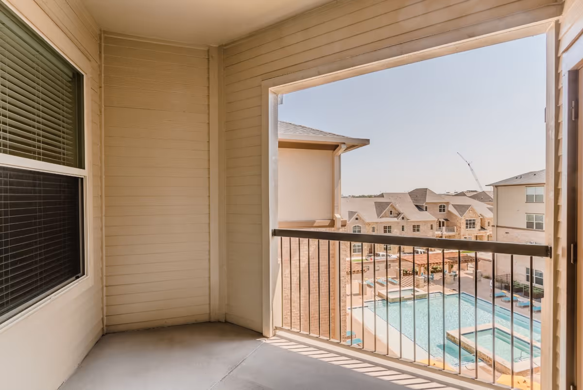 View from a covered balcony with beige walls and a railing overlooking a swimming pool and surrounding buildings under a clear sky.