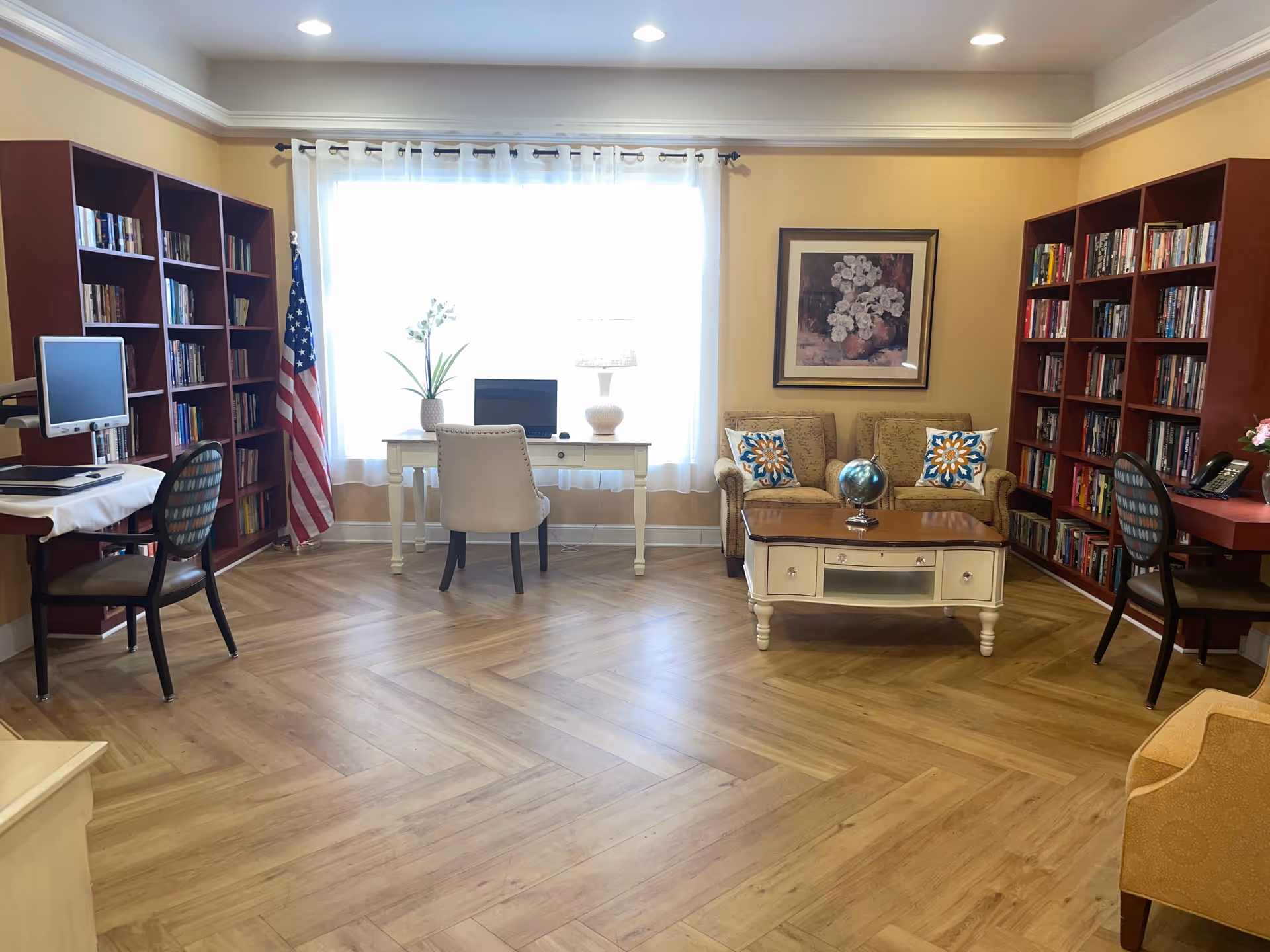 Bright communal lounge/library with bookshelves, a desk and computer by a large window, and a sofa with a coffee table.